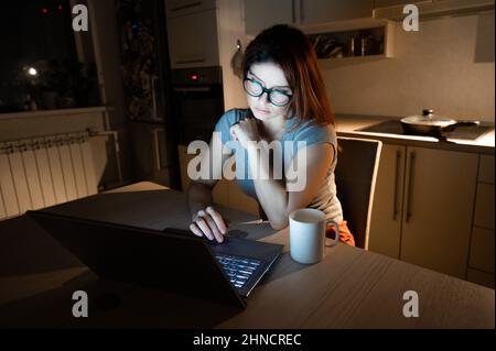Caucasian woman wearing glasses at a wireless computer at night in the kitchen. Girl works overtime in the home office. Freelancer is typing on a Stock Photo