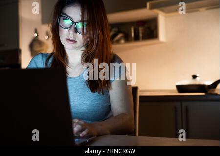 Caucasian woman wearing glasses at a wireless computer at night in the kitchen. Girl works overtime in the home office. Freelancer is typing on a Stock Photo