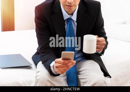 Shot of a handsome businessman sitting on the bed with smart phone and coffee Stock Photo