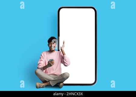 Excited indian guy sitting by huge smartphone with blank screen Stock Photo
