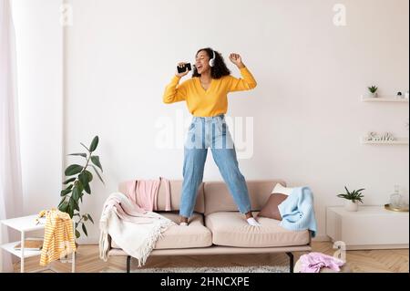 Cool black woman in headphones using smartphone as mic, standing on couch, singing songs, checking mobile music app Stock Photo