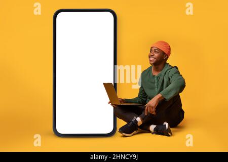 African american man sitting with laptop pc near big cellphone with empty white screen, presenting cool app or website Stock Photo