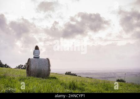 Back view of cropped unrecognizable female sitting on hay roll on grassy field in countryside against cloudy sky Stock Photo