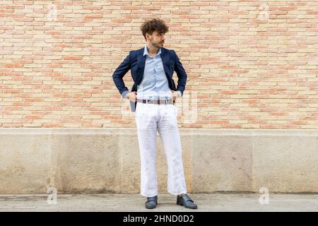 Handsome bearded Hispanic male in formal suit looking away while standing near brick wall on street Stock Photo
