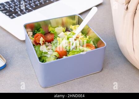 Salad with fresh green lettuce and cherry tomatoes served in square metal container with plastic spoon placed near modern netbook Stock Photo