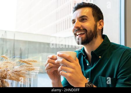 Side view of cheerful bearded male entrepreneur drinking hot coffee while sitting at table with slice of cake in light modern cafeteria during lunch b Stock Photo