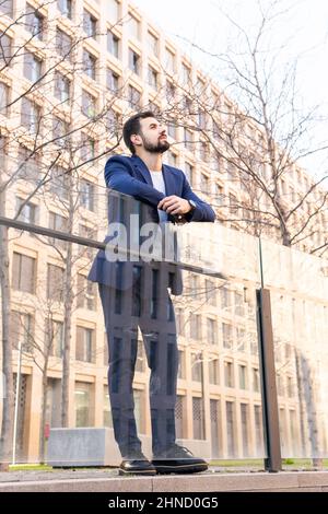 From below thoughtful bearded male entrepreneur in elegant wear standing near modern building leaning on glass wall on street of city with trees Stock Photo