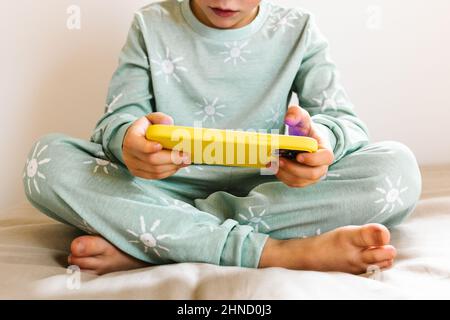 Cropped unrecognizable boy in sleepwear playing game on cellphone while sitting on bed near window in light bedroom Stock Photo