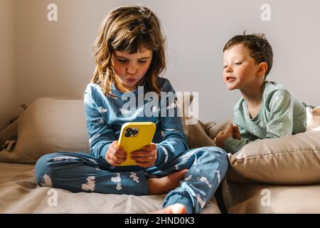 Siblings in sleepwear playing game on modern cellphone while sitting together on comfortable bed in light bedroom on morning time Stock Photo