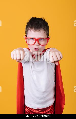 Isolated boy in glasses and red superhero costume looking at camera while pretending to fly on yellow background in studio Stock Photo