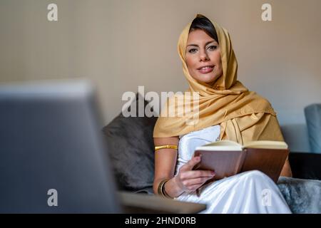 Muslim Woman in white dress and golden headscarf smiling and looking at camera while sitting on sofa near laptop and checking data in notebook during Stock Photo