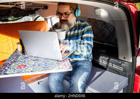 Crop mature bearded male in headphones watching video on netbook while sitting in mini camper during road trip Stock Photo