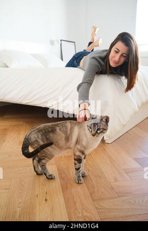 Content female owner playing with cute gray cat while lying on comfortable bed in light bedroom with wooden flooring Stock Photo