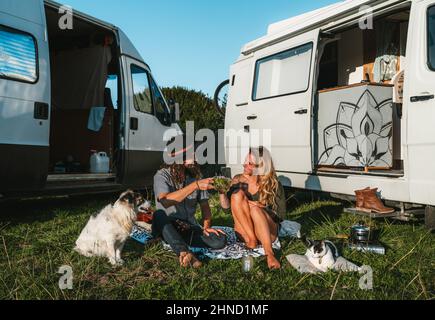 Full body of positive traveling couple looking at each other and clinking glasses with tea while sitting with dog and cat near camping vans during roa Stock Photo