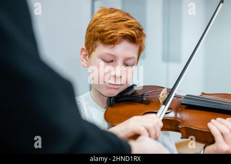 Crop music teacher standing near redhead boy with violin in classroom Stock Photo