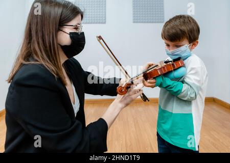 Female music teacher in mask sitting with bow near boy with violin in classroom Stock Photo