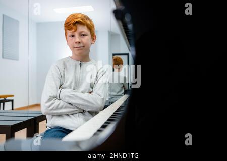 Content ginger preteen child sitting near piano in classroom and looking at camera Stock Photo