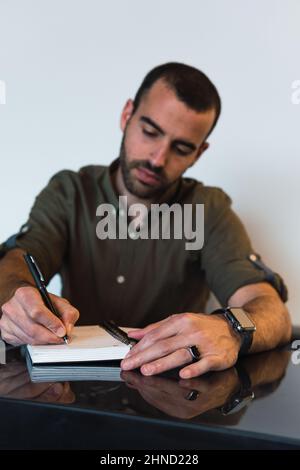 Focused male taking notes in notepad while sitting at table in light room during work time Stock Photo