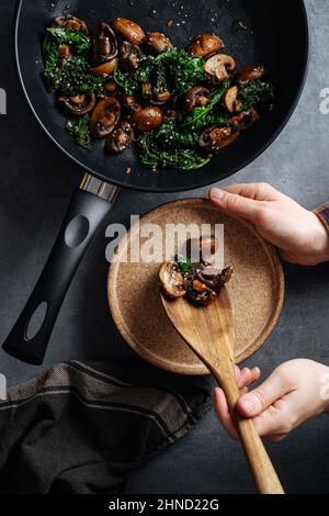 From above of crop anonymous person with wooden spatula putting tasty sauteed mushrooms with greens from frying pan on plate Stock Photo