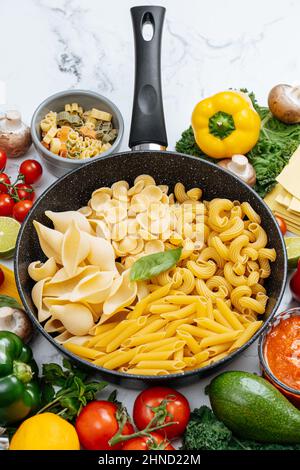 From above of frying pan with various uncooked pasta surrounded with assorted raw vegetables on marble table in light kitchen Stock Photo