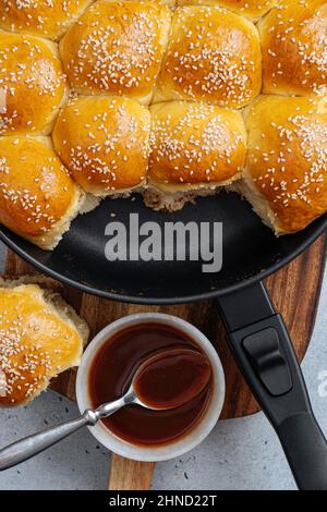 From above of tasty pull apart bread rolls with sesame seeds in frying pan served on wooden cutting board with sweet sauce Stock Photo