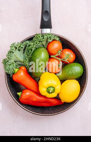 Top view of frying pan with raw ripe peppers and avocados near cherry tomatoes and greens on light background in kitchen Stock Photo