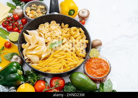 From above of frying pan with various uncooked pasta surrounded with assorted raw vegetables on marble table in light kitchen Stock Photo