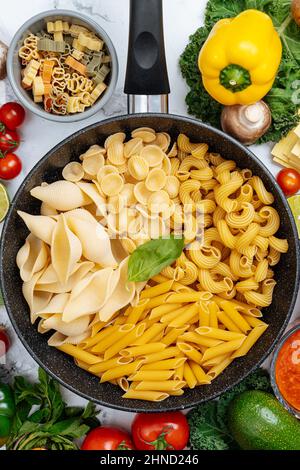 From above of frying pan with various uncooked pasta surrounded with assorted raw vegetables on marble table in light kitchen Stock Photo