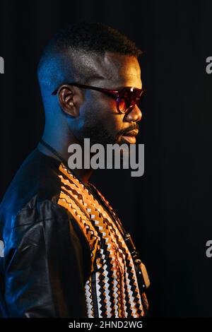 Side view of serious bearded man with african outfit in sunglasses looking away while standing on black background Stock Photo