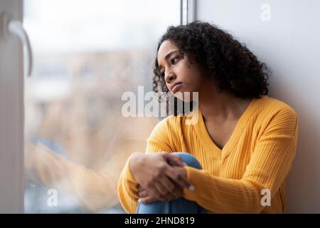 Depressed young African American woman sitting near window, looking outside on street, feeling hopeless at home