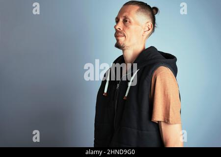 A close-up photo of a young man in a vest on the side. A man with long hair gathered in a knot or a man's bun. on a blue background, men's hairstyles, Stock Photo