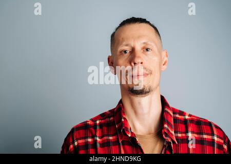 A close-up photo of a young man in a red plaid shirt. A man with long hair gathered in a knot or a man's bun looks at the camera. on a blue background Stock Photo