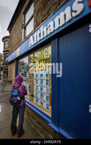 Ramsbottom, Lancashire, UK, Wednesday February 16, 2022. The start of Storm Dudley hits the North West of England on Bridge Street, Ramsbottom, Lancashire. A shopper stops to check out the summer sun offers outside this travel agent. Credit: Paul Heyes/Alamy News Live Stock Photo