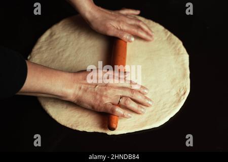 The chef's hands roll out fresh homemade pizza dough on the kitchen table with a rolling pin, Beautiful female hands roll out the dough, make pasta, r Stock Photo