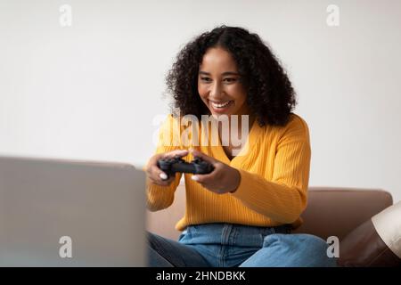 Cool young black woman with gaming joystick playing computer game on laptop pc at home Stock Photo