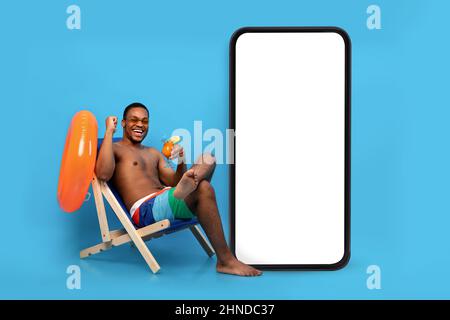 Excited black guy with tropical cocktail chilling in lounge chair near huge smartphone with blank screen for your ad Stock Photo