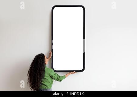 Woman holding huge smartphone with empty screen for your advertisement on white wall, free space, mockup Stock Photo