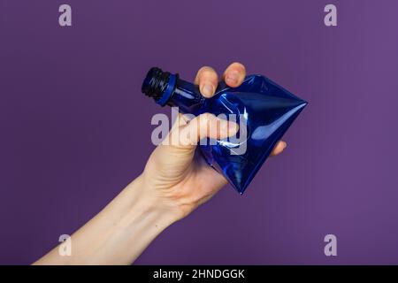 Woman's hand holds a crumpled blue plastic bottle on a purple background Stock Photo