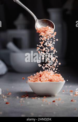 Himalayan pink salt in crystals is poured into a small white bowl. Stock Photo