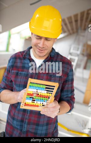 Construction worker with shirt and yellow helm calculates with abacus calculator on construction site in a loft Stock Photo