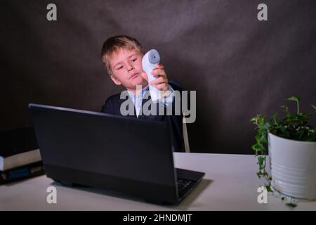 A happy boy in a school suit with a thermometer at the computer, copy space on a dark studio background Stock Photo