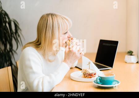 Mature adult woman sitting in cafe with coffee mug eating sandwiches and working online at laptop. Businesswoman in typing on notebook computer, in Stock Photo