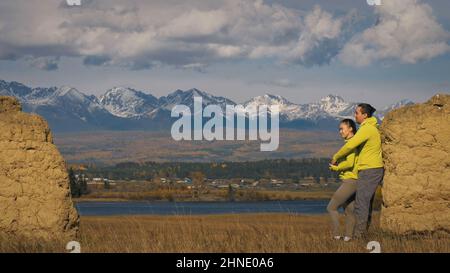 Man and woman in yellow green sportswear. Lovely couple of travelers hug and kiss near old stone enjoying highland landscape. Two travelers are walking against the backdrop of snow-capped mountains. Stock Photo