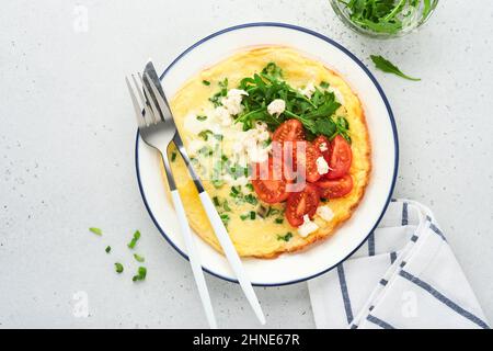 Breakfast fried eggs. Rustic omelette or frittatas with green onions, cheese mozzarella, green arugula and tomatoes on light stone background. Healthy Stock Photo