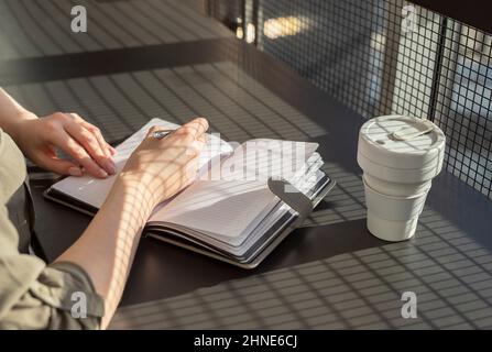 Hands closeup taking notes in planner. Woman sitting at desk with eco cup in cafe and writing plans, thoughts in diary. High quality photo Stock Photo