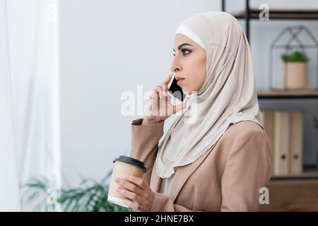 side view of muslim woman talking on smartphone while standing with coffee to go at home Stock Photo