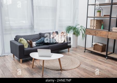 happy muslim woman listening music in headphones while lying on couch in living room Stock Photo