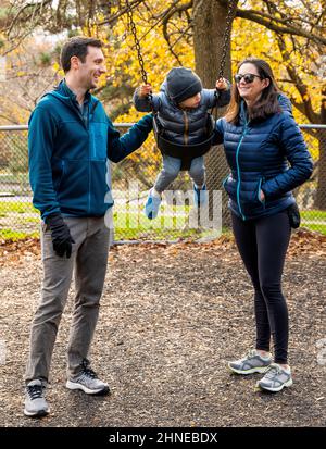 Young parents pushing two-year-old boy on a city playground swing; Philadelphia; Pennsylvania; USA Stock Photo