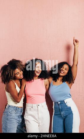 Three beautiful female friends in bright casuals having fun while leaning pink wall Stock Photo