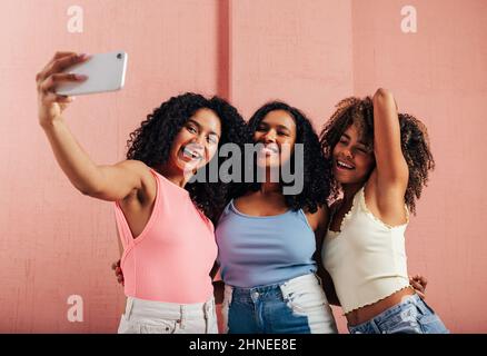 Three happy women with curly hair taking selfie against pink wall Stock Photo